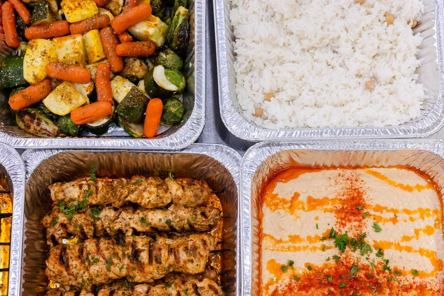 Overhead view of catering trays with roasted vegetables, chickpea rice, chicken skewers and hummus